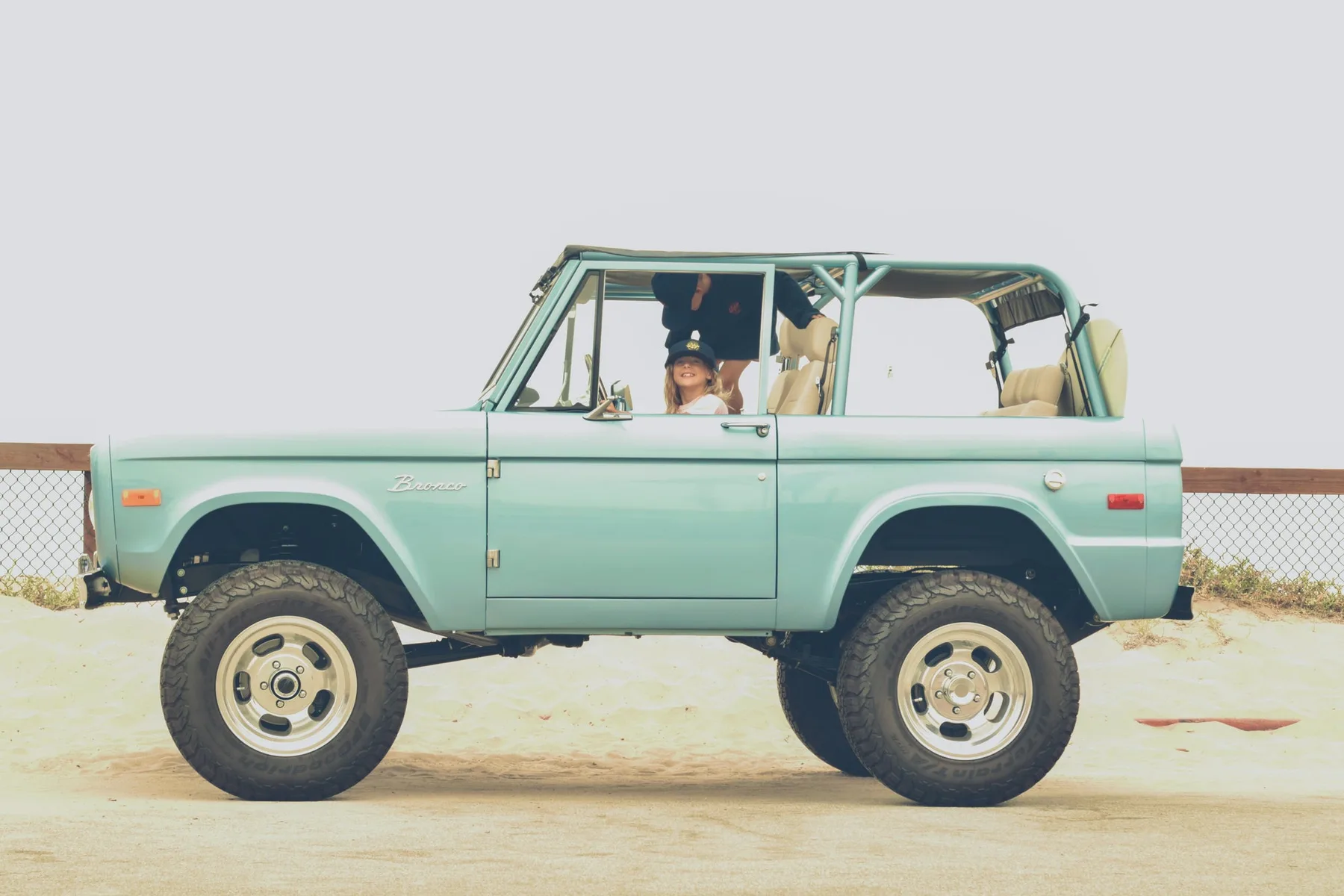 Lily's seafoam 1970s Ford Bronco parked on the sand with the top off and a child sitting in the driver's seat
