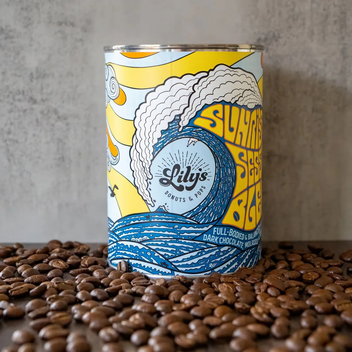 A Lily's Sunrise Session Blend coffee can, illustrated with a breaking wave and sunrise, standing in a spill of roasted beans