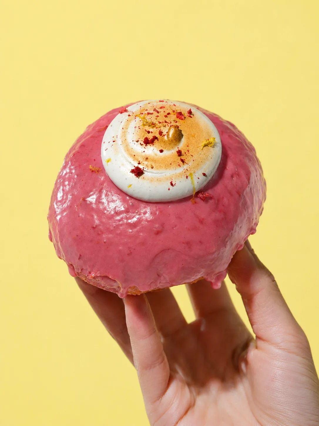 A raised donut with glossy pink raspberry glaze and a torched meringue swirl, held up against a butter-yellow wall