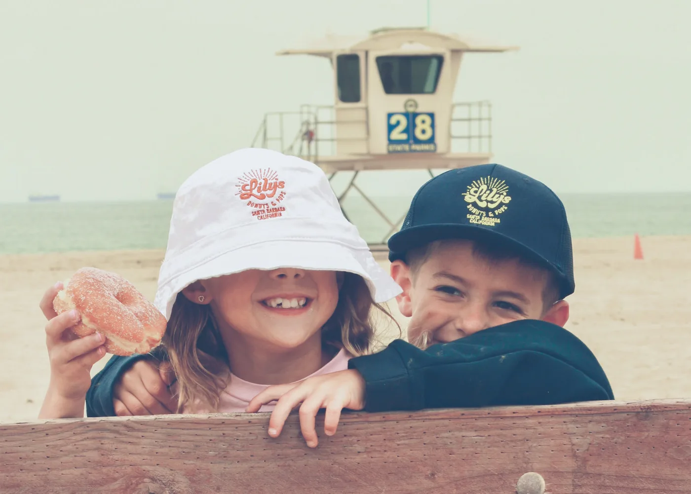Two children in Lily's caps grinning over a fence at the beach, one holding a sugared donut, lifeguard tower 28 behind them