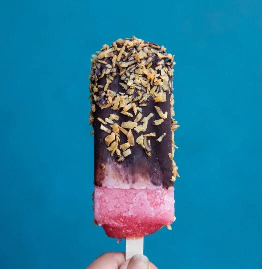 A strawberry popsicle dipped in dark chocolate and rolled in toasted coconut, held against a teal wall